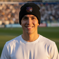 A smiling man wearing a black knit beanie with a Tigres de Quintana Roo logo and a white t-shirt, standing in a stadium with blurred spectators in the background.