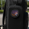 A black tactical-style backpack with a front pocket featuring a circular embroidered patch that reads 'The Tigers' with a tiger head and orange stars, photographed outdoors on a paved surface with blurred greenery and buildings in the background.