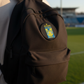 A black backpack with a circular Tigres UANL crest patch on the front, worn by a person on a green soccer field at sunset.
