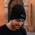A man wearing a black knit beanie with a white embroidered Tlaxcala logo, standing outdoors in front of a warm-toned stone archway and orange wall.