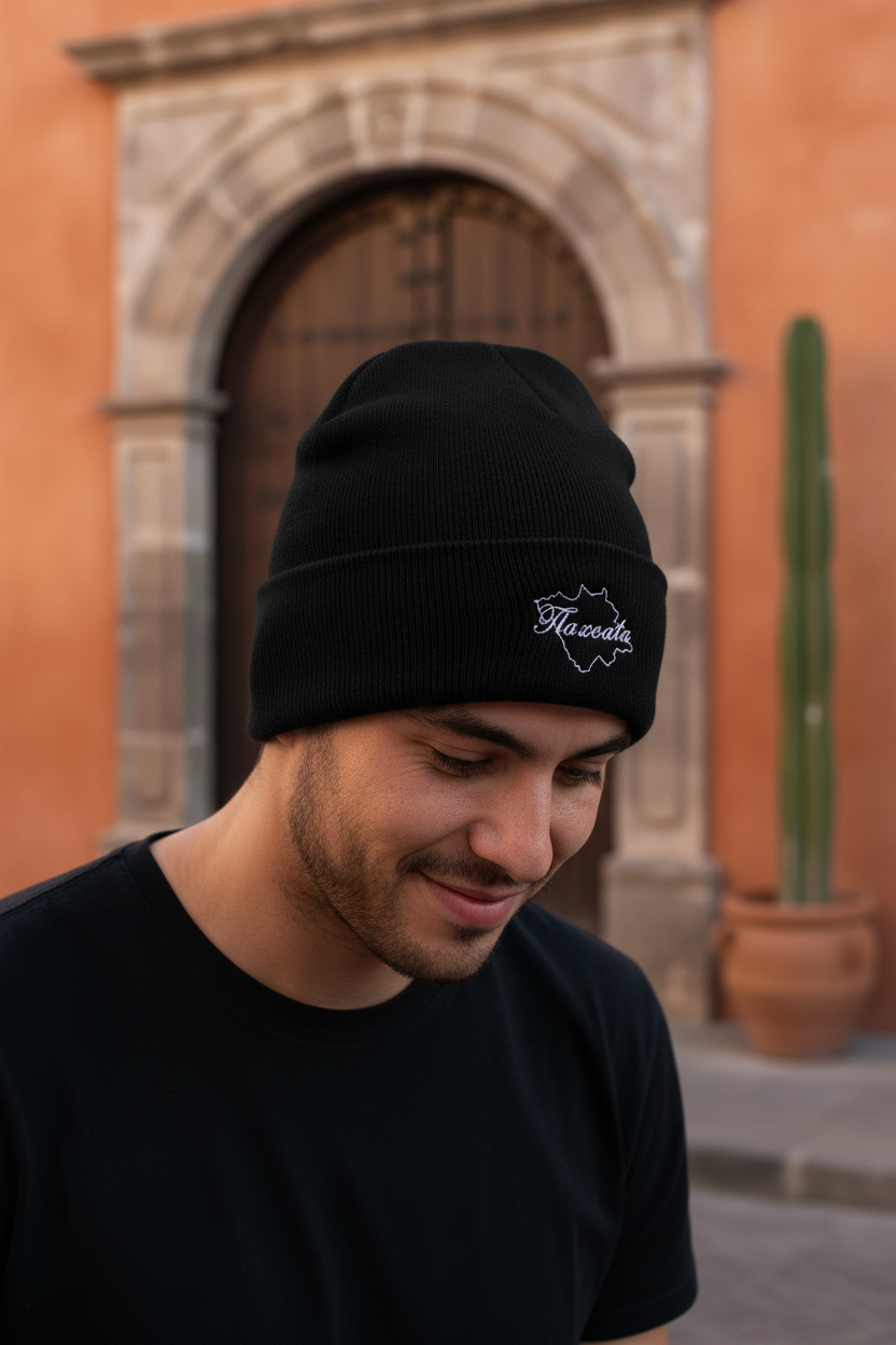 A man wearing a black knit beanie with a white embroidered Tlaxcala logo, standing outdoors in front of a warm-toned stone archway and orange wall.