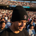 A man wearing a black Toros de Tijuana knit beanie with a circular embroidered logo on the front, seated in a crowded stadium during warm golden hour sunlight.
