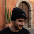 A man wearing a black knit beanie with a small white embroidered logo, standing outdoors in front of a warm terracotta wall and stone archway.
