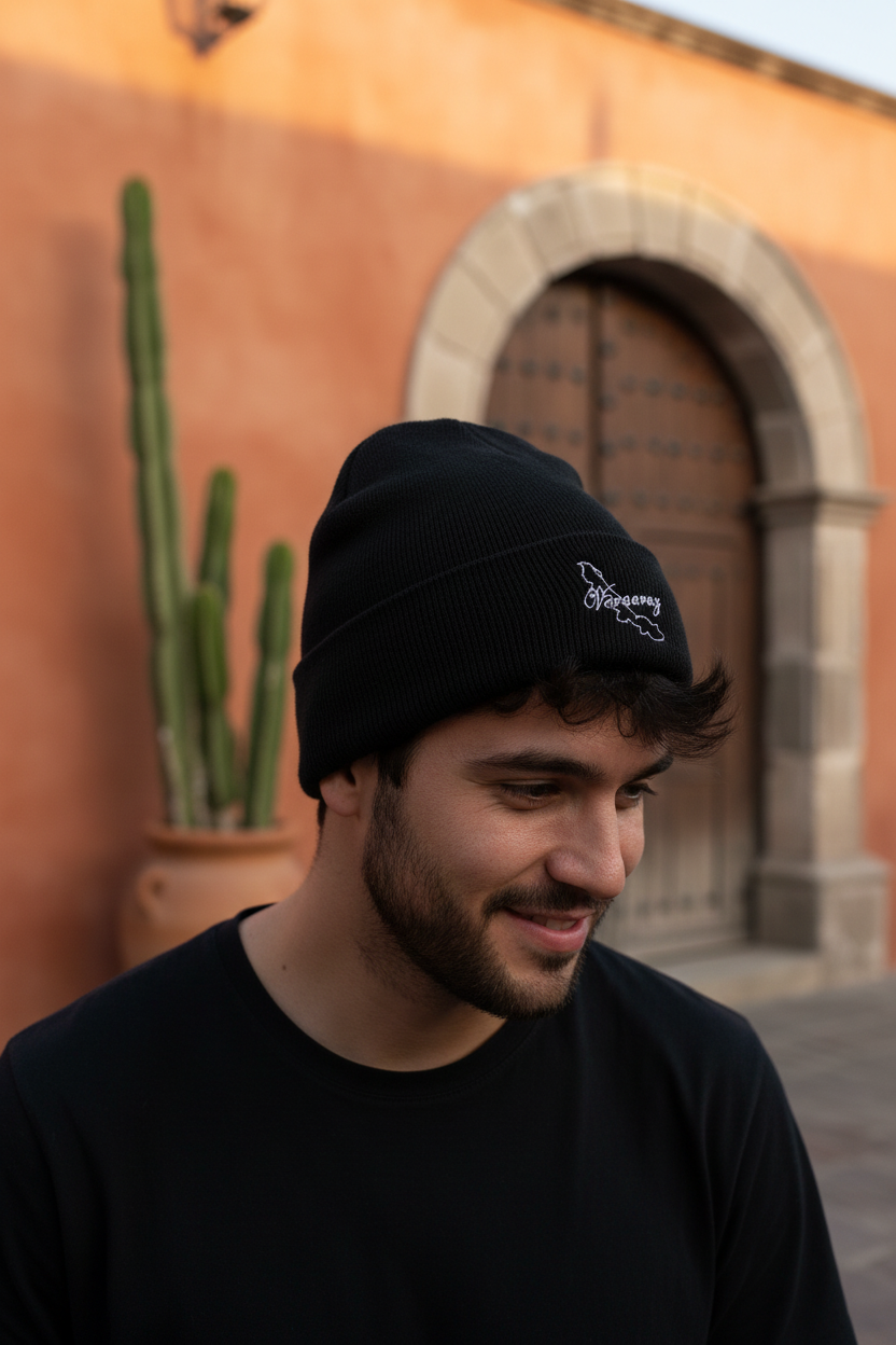 A man wearing a black knit beanie with a small white embroidered logo, standing outdoors in front of a warm terracotta wall and stone archway.