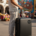 A man in a white t-shirt and light gray pants stands in a colorful outdoor plaza next to a black hard-shell rolling suitcase with a telescoping handle extended.