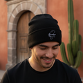 A man wearing a black knit beanie with a white embroidered Zacatecas logo, standing outdoors in front of a warm-toned stucco wall and cactus.