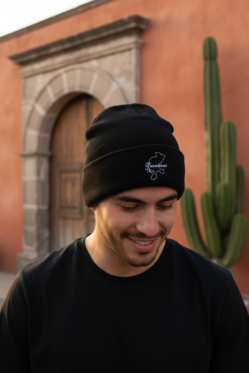 A man wearing a black knit beanie with a white embroidered Zacatecas logo, standing outdoors in front of a warm-toned stucco wall and cactus.