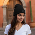 A woman wearing a black knit beanie with a small white embroidered logo, standing outdoors in front of a warm-toned arched wooden door.