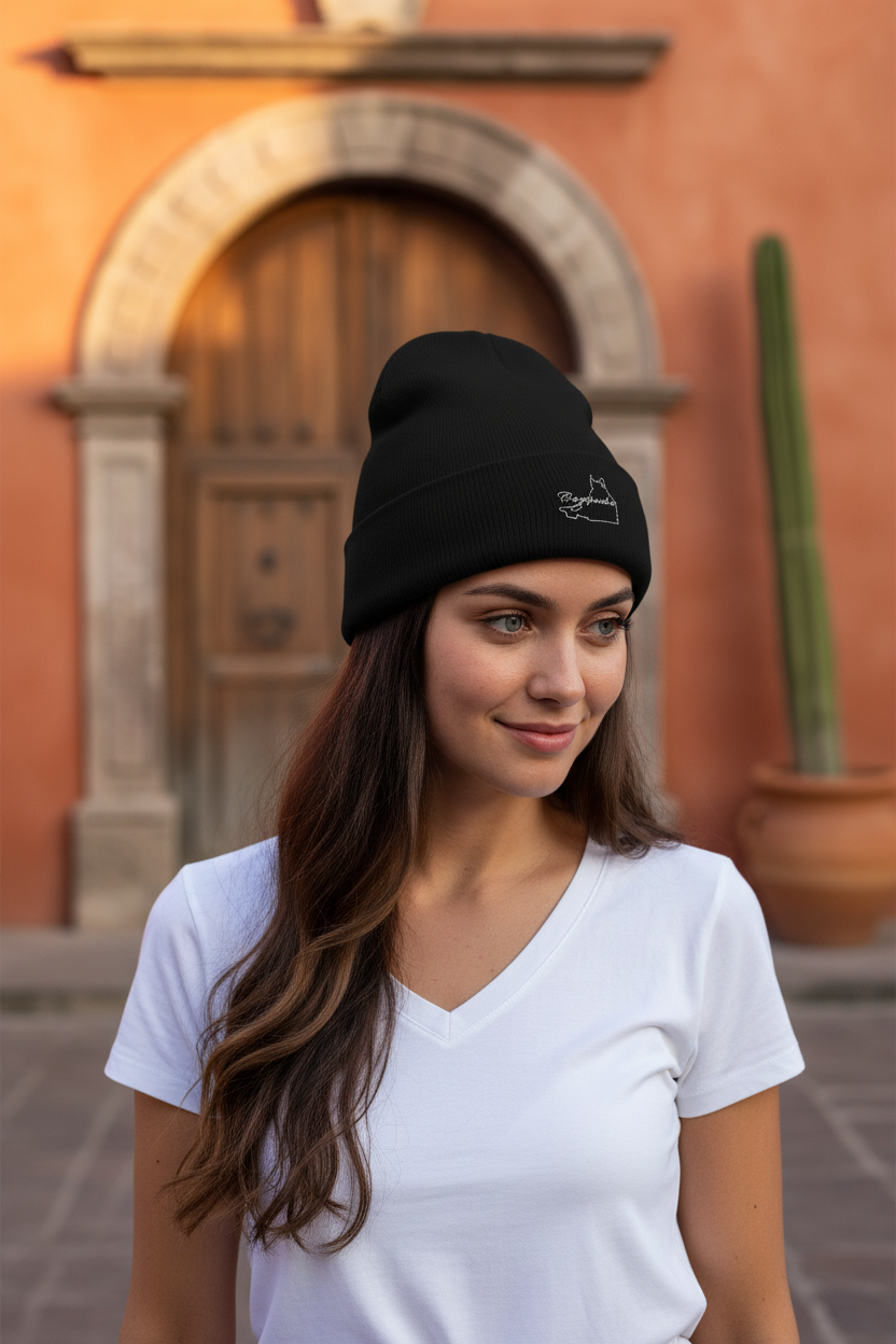 A woman wearing a black knit beanie with a small white embroidered logo, standing outdoors in front of a warm-toned arched wooden door.
