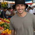 A man wearing a black bucket hat with a small white graphic logo and a gray t-shirt, standing at an outdoor market with produce displays in the background.