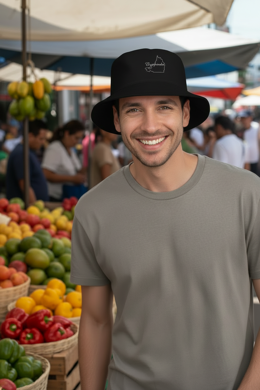 A man wearing a black bucket hat with a small white graphic logo and a gray t-shirt, standing at an outdoor market with produce displays in the background.