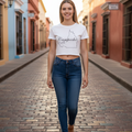 A woman walking down a cobblestone street wearing a white crop top with a black outline map of Cape Town and the word 'Campeche printed on the front, paired with dark blue jeans and brown sandals.