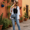 A woman stands on a stone walkway in front of a warm orange stucco building, wearing a white open-front cardigan over a black top, light blue jeans, and black slide sandals. She is holding a drink and wearing a blue crossbody bag.