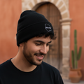 A man wearing a black knit beanie with a small white embroidered Chihuahua logo, standing outdoors in front of a warm terracotta wall and stone archway.