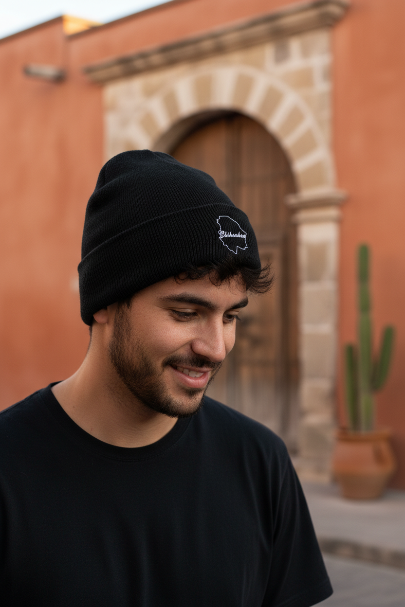 A man wearing a black knit beanie with a small white embroidered Chihuahua logo, standing outdoors in front of a warm terracotta wall and stone archway.
