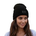 A woman with long brown hair wearing a  Chihuahua black knit beanie with a small white outline logo on the front, paired with a white V-neck t-shirt, set against a plain white background.
