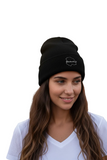 A woman with long brown hair wearing a  Chihuahua black knit beanie with a small white outline logo on the front, paired with a white V-neck t-shirt, set against a plain white background.