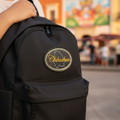 A black backpack with a circular embroidered patch featuring the outline of Chihuahua and the word 'Chihuahua' in gold script, shown being worn by a person in an outdoor setting with a blurred mural in the background.