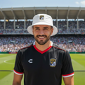A smiling man wearing a white bucket hat and a black soccer jersey with red and white trim stands on a stadium field with a large stadium in the background.