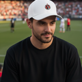 A man wearing a white baseball cap with a circular red and black Club Tijuana logo and a black t-shirt, seated in a stadium with a soccer field and crowd in the background.