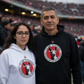 A man and a woman standing together in a stadium, both wearing matching Club Tijuana hoodies. The woman wears a white hoodie, and the man wears a black hoodie, both featuring the club's circular logo on the chest.
