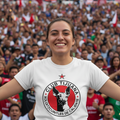 A smiling woman in a crowded stadium wearing a white Club Tijuana graphic t-shirt with a red and black circular logo on the chest.