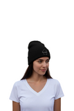 A woman wearing a black knit beanie with a small white embroidered logo on the front, paired with a white V-neck t-shirt, set against a plain white background.