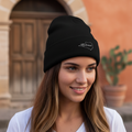 A woman wearing a black knit beanie with a small white embroidered logo, standing outdoors in front of a warm-toned stucco building with a wooden door.