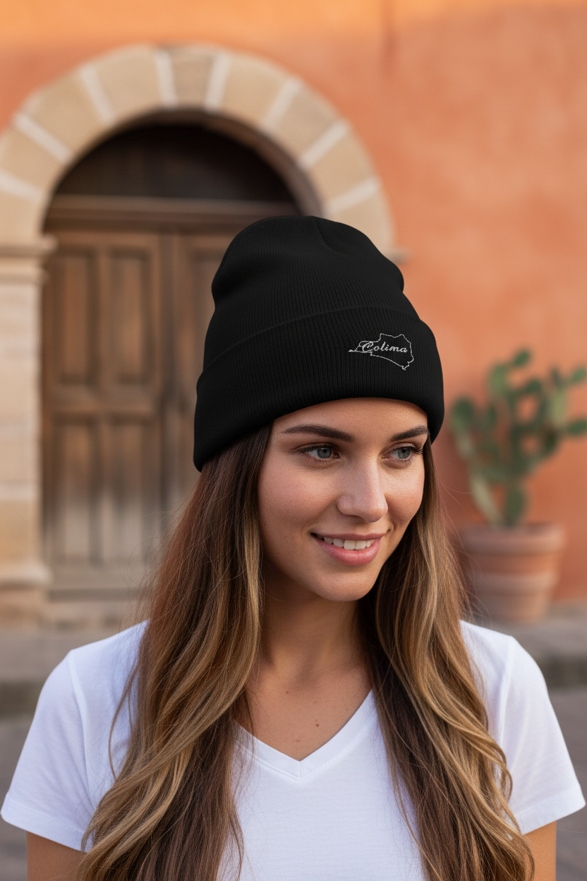 A woman wearing a black knit beanie with a small white embroidered logo, standing outdoors in front of a warm-toned stucco building with a wooden door.