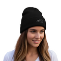 A woman with long brown hair wearing a black knit beanie with a white embroidered Colima logo, paired with a white t-shirt, set against a solid white background.