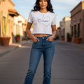 A woman stands on a sunlit street wearing a white short-sleeve crop top with a black script outline graphic and blue jeans.