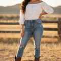 A woman stands outdoors in a dry field wearing a white off-the-shoulder blouse, light-wash distressed blue jeans, and brown cowboy boots.