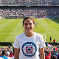 A smiling woman stands in a crowded soccer stadium wearing a white Cruz Azul graphic T-shirt. The background shows a green field, packed stands, and a large scoreboard.