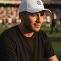 A man wearing a white Cruz Azul baseball cap with a circular crest and a black t-shirt, sitting in a stadium during golden hour.