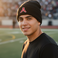A smiling young man wearing a black Acereros de Monclova knit beanie with a red and white 'A' logo on the front, standing on a football field at sunset.