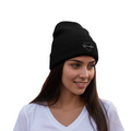 A woman with long dark hair wearing a black Durango knit beanie with a small white embroidered logo on the side, paired with a white V-neck t-shirt, set against a plain white background.