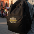 A black nylon backpack with a circular gold and white 'Durango' logo patch on the front pocket, worn by a person in a sunny outdoor street setting.