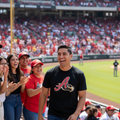 A man in a black Aquila de Veracruz  t-shirt with a red and gold logo stands in a crowded baseball stadium, smiling and clapping along with fans wearing red and black apparel.