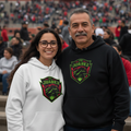 A man and a woman standing together in stadium seating, both wearing matching black and white hoodies with a green and red 'FC Juarez' shield logo on the chest.