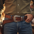 Close-up of a person wearing a brown western-style leather belt with a large engraved silver buckle, paired with blue jeans and an olive green button-up shirt.