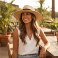 A woman sitting at an outdoor wooden table wearing a straw wide-brim hat, a white sleeveless top, and denim shorts in a warm, sunlit patio setting.