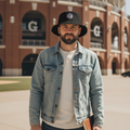 A man wearing a light-wash denim jacket, dark jeans, a black Generales de Durango bucket hat, and a white t-shirt stands in front of a large brick stadium with 'G' lettering on the facade.