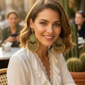 A woman with wavy brown hair wearing large, ornate gold-toned circular drop earrings and a white wrap-style blouse, seated at an outdoor cafe table with warm sunlight and blurred background figures.