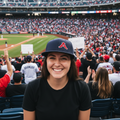 A smiling woman in a baseball stadium wearing a black t-shirt and a Acereros de Monvlova navy blue cap with a red letter 'A' logo, with a crowded stadium and baseball field in the background.