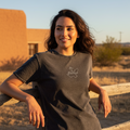 A woman wearing a dark charcoal gray t-shirt with a white script logo on the left chest, leaning against a wooden fence in a desert setting with a tan adobe building in the background.
