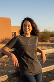 A woman wearing a dark charcoal gray t-shirt with a white script logo on the left chest, leaning against a wooden fence in a desert setting with a tan adobe building in the background.