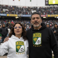 A man and a woman standing together in a stadium, both wearing hoodies with the Leon soccer team crest. The woman wears a white hoodie, and the man wears a black hoodie.