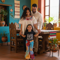 A family of three stands together in a brightly lit, colorful home interior. The young girl in the center wears a black graphic T-shirt that reads 'take a little DIRT BOOTS' and matching tie-dye bell-bottom pants.