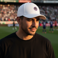 A man wearing a white baseball cap with a circular Mazatlan FC logo and a black t-shirt, standing on a soccer field with a stadium crowd in the background.