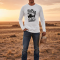A man stands in a dry grassy field at sunset wearing a white long-sleeve graphic shirt with a buffalo print, blue jeans, and holding a cowboy hat.